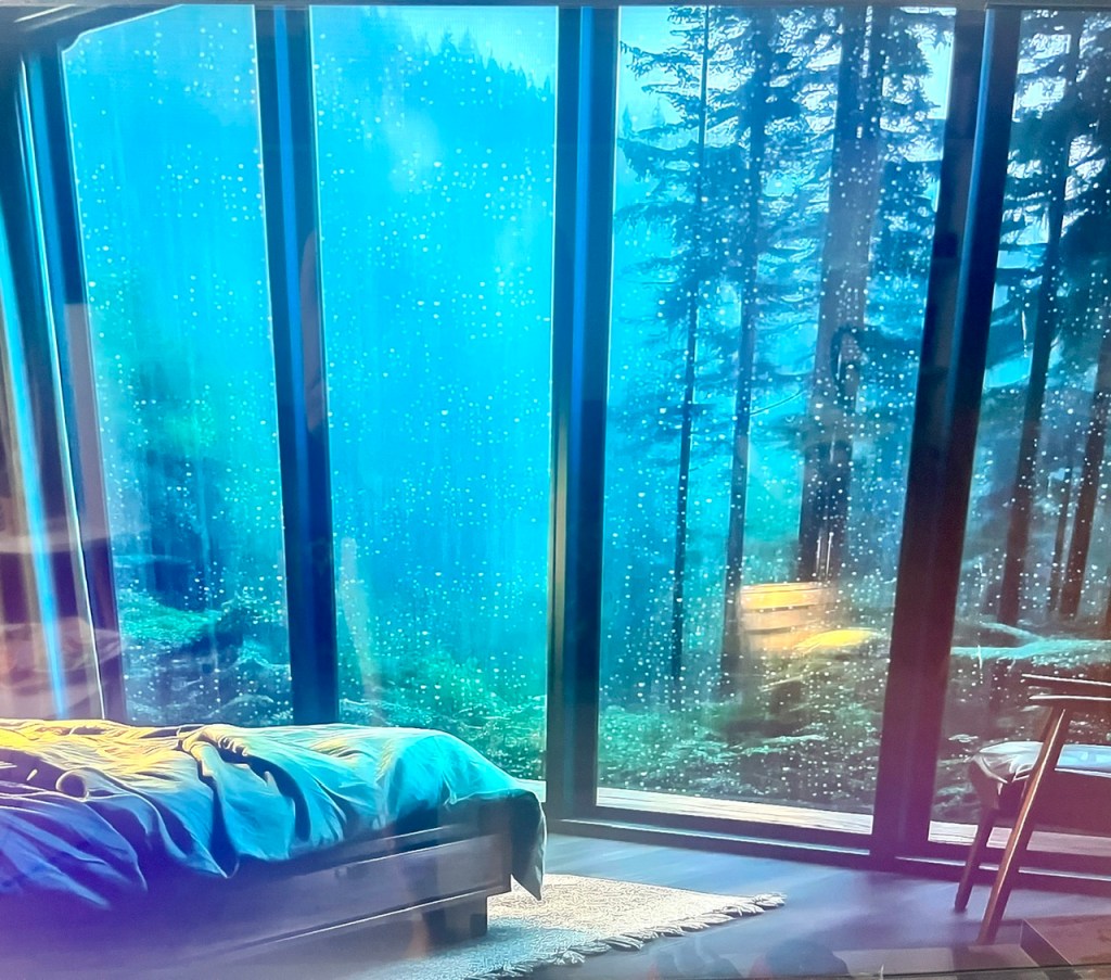 Một góc phòng ngủ với giường và một chiếc ghế, bên ngoài có khung cửa kính lớn nhìn ra rừng cây xanh mát, trời đang mưa.